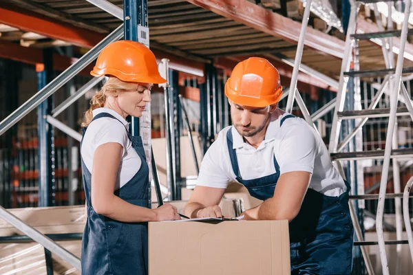 Concentrated warehouse workers looking at clipboard while standing near cardboard box — Stock Photo