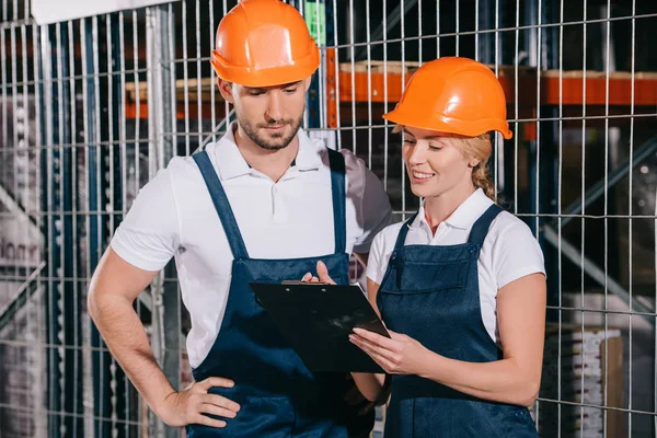 Lächelnde Arbeiterin hält Klemmbrett, während sie neben konzentriertem Arbeiter steht — Stockfoto