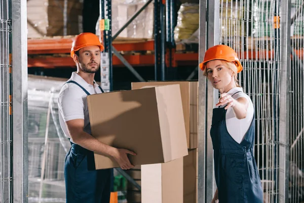 Trabajador de almacén que sostiene la caja de cartón cerca de workwoman que señala con la mano - foto de stock