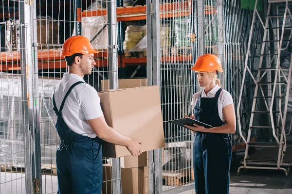 Cargador que sostiene la caja de cartón cerca de workwoman que escribe en portapapeles - foto de stock