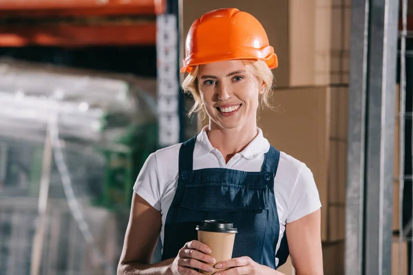 Hermosa trabajadora en casco sosteniendo café para ir y sonriendo a la cámara - foto de stock