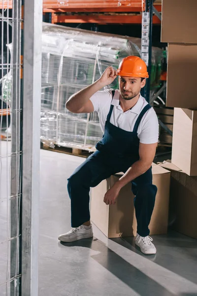 Tired loader sitting on cardboard box in wearhouse and touching helmet — Stock Photo