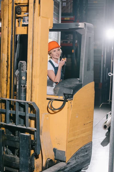 Trabajadora sonriente sentada en el cargador de carretillas elevadoras, hablando en walkie talkie y mirando hacia otro lado - foto de stock