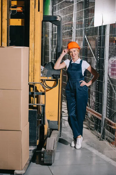 Trabalhadora alegre de pé perto carregador de empilhadeira com a mão no quadril, tocando capacete e olhando para a câmera — Fotografia de Stock