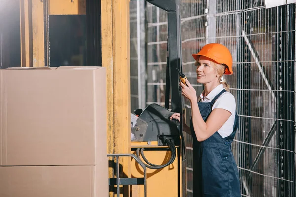 Smiling workwoman talking on walkie talkie while standing near forklift loader — Stock Photo