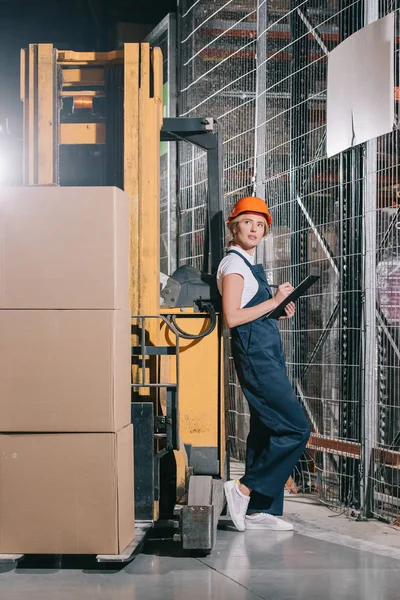 Atractiva trabajadora escribiendo en el portapapeles y mirando hacia otro lado mientras está de pie cerca de la cargadora de carretillas elevadoras - foto de stock