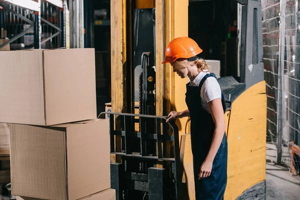 Workwoman in overalls and helmet standing near foklift loader in warehouse — Stock Photo
