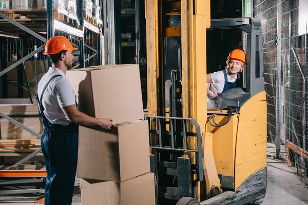 Lächelnde Arbeiterin sitzt im Gabelstapler und schaut Arbeiter mit Pappschachtel an — Stockfoto