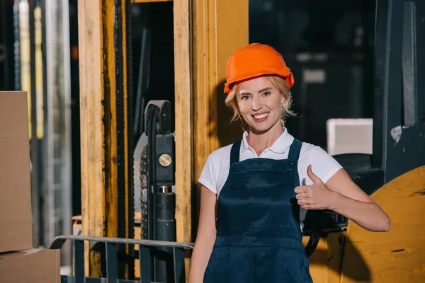 Trabajadora alegre mirando a la cámara y mostrando el pulgar hacia arriba mientras está de pie cerca de la carretilla elevadora - foto de stock