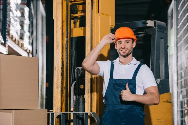 Sorrindo trabalhador armazém tocando capacete e mostrando polegar para cima enquanto está perto de carregador de empilhadeira — Fotografia de Stock