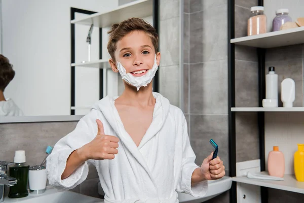 Menino feliz com espuma de barbear no rosto segurando navalha e mostrando o polegar para cima — Fotografia de Stock