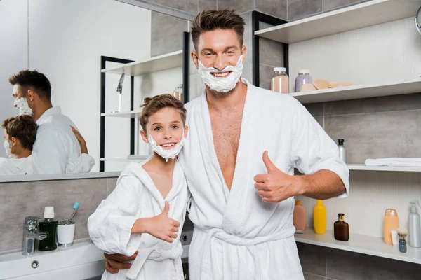 Cheerful father and son with shaving foam on faces showing thumbs up in bathroom — Stock Photo