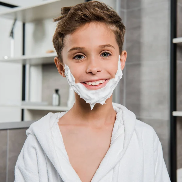 Menino alegre com espuma de barbear no rosto sorrindo no banheiro — Fotografia de Stock