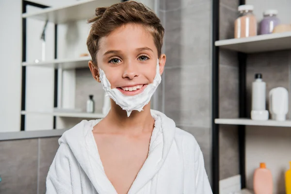 Garçon heureux avec mousse à raser sur le visage souriant dans la salle de bain — Photo de stock