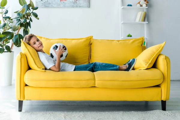 Happy boy in white t-shirt lying on yellow sofa with football — Stock Photo