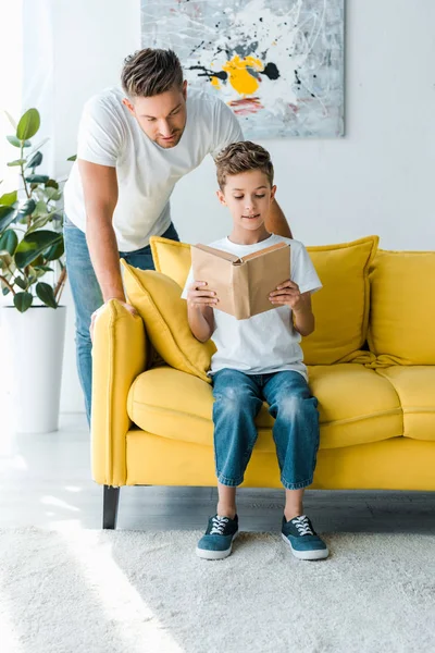 Schöner Vater, der neben Sohn steht und Buch liest, während er auf dem Sofa sitzt — Stock Photo