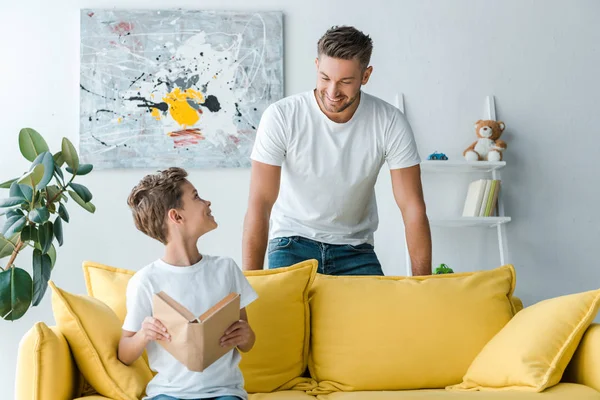 Happy father standing near son holding book while sitting on sofa — Stock Photo