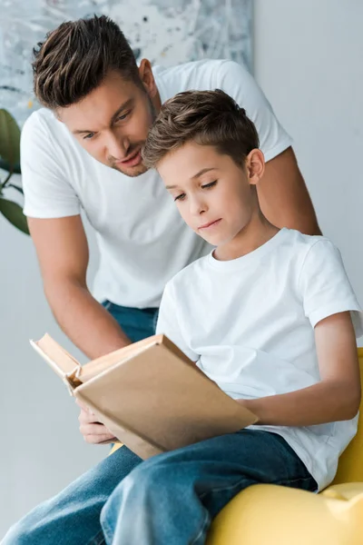 Selective focus of father standing near son holding book while sitting on sofa — Stock Photo