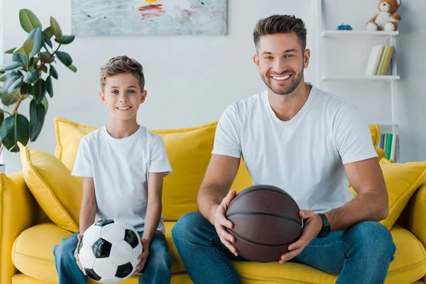 Happy man holding basketball near son with football at home — Stock Photo