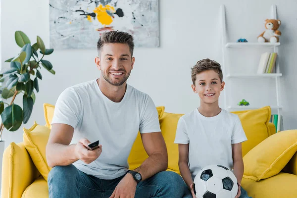 Alegre padre sosteniendo mando a distancia cerca de hijo con fútbol sentado en el sofá - foto de stock