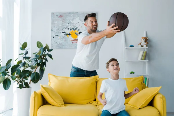 Happy father holding basketball above head of son sitting on sofa — Stock Photo