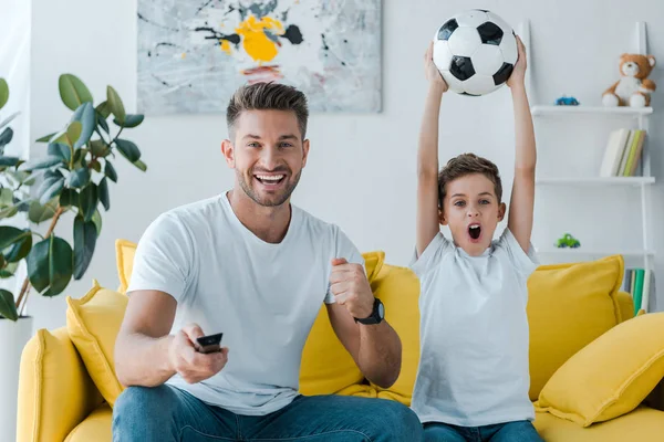 Pai alegre segurando controlador remoto perto do filho com futebol acima da cabeça — Fotografia de Stock