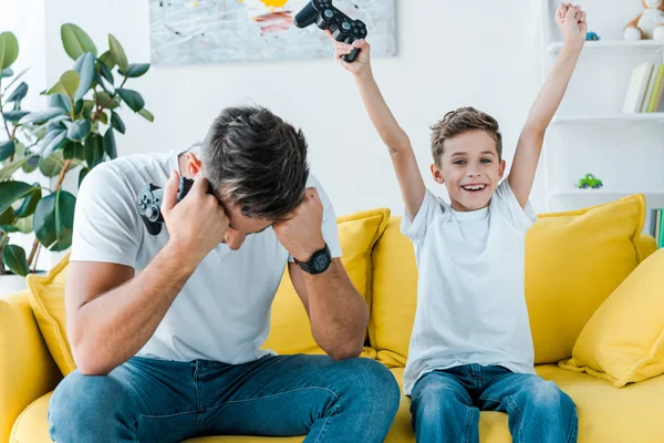 KYIV, UCRANIA - 2 DE OCTUBRE DE 2019: padre molesto cerca de hijo feliz celebrando el triunfo mientras sostiene gamepad - foto de stock