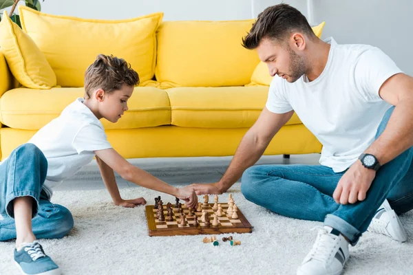 Vista lateral de padre e hijo guapos jugando ajedrez en la alfombra - foto de stock