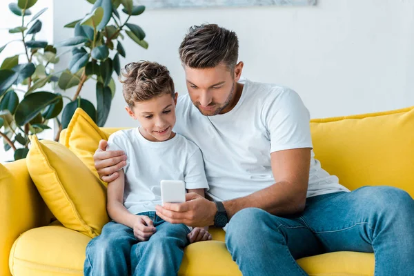 Cute son and father looking at smartphone while sitting on sofa — Stock Photo