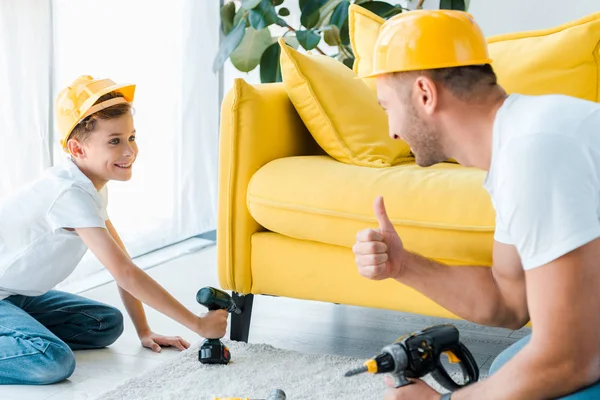 Foco seletivo da criança feliz no capacete de segurança segurando martelo broca e olhando para o pai mostrando polegar para cima — Fotografia de Stock