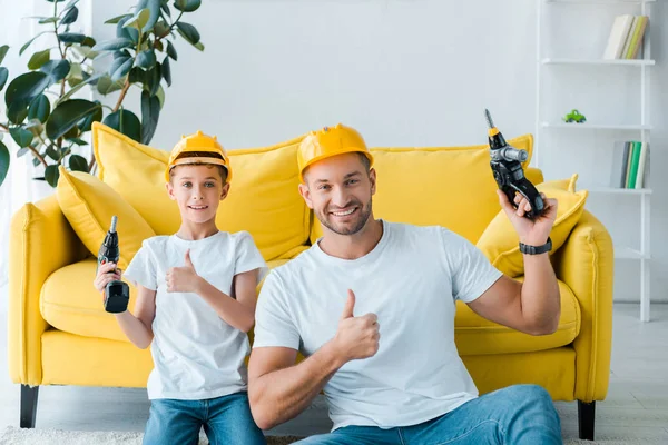 Happy son and handsome father in safety helmets showing thumbs up and holding hammer drills — Stock Photo