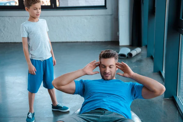 Lindo hijo de pie cerca de padre haciendo ejercicio en la pelota de fitness - foto de stock