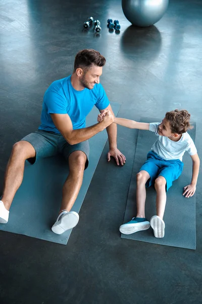 Vue aérienne du père et du fils heureux tenant la main tout en étant assis sur des tapis de fitness — Photo de stock