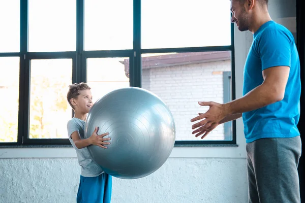 Carino bambino tenendo palla fitness e guardando il padre in palestra — Foto stock