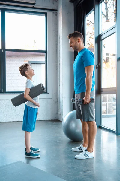 Vista lateral del niño mirando a padre mientras que está parado con la estera de la aptitud en gimnasio — Stock Photo