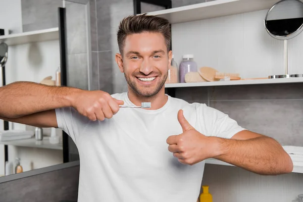 Hombre alegre en camiseta blanca sosteniendo el cepillo de dientes y mostrando el pulgar hacia arriba en el baño - foto de stock