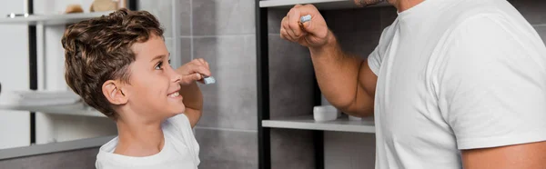 Plano panorámico de niño sosteniendo cepillo de dientes y mirando a padre en baño - foto de stock