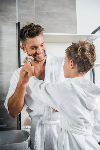 Избирательный фокус счастливого отца глядя на сына, держащего кисть для бритья — стоковое фото