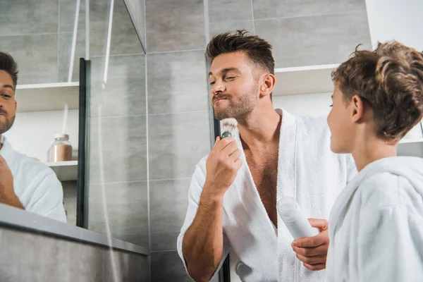 Enfoque selectivo de padre guapo sosteniendo cepillo de afeitar cerca de la cara mientras está de pie cerca de hijo en el baño - foto de stock