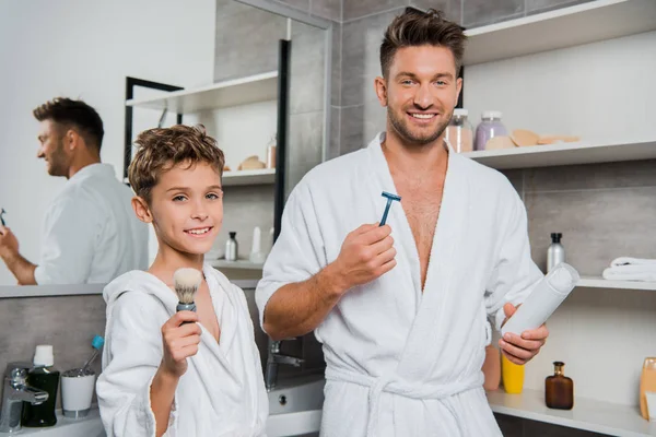 Feliz padre sosteniendo la navaja y puede con espuma de afeitar cerca de hijo con cepillo de afeitar - foto de stock