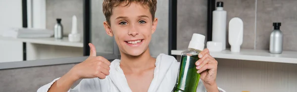 Plan panoramique de garçon heureux tenant bouteille avec bain de bouche vert et montrant pouce dans la salle de bain — Photo de stock