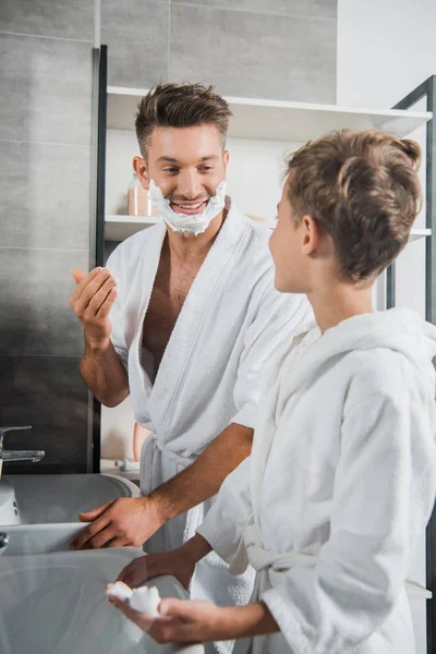 Enfoque selectivo de padre alegre con espuma de afeitar en la cara mirando hijo en el baño - foto de stock