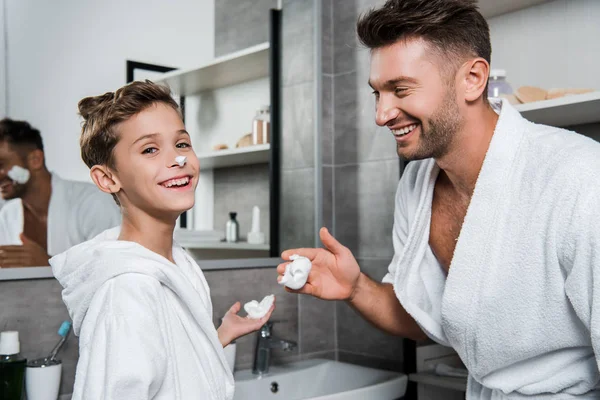 Niño feliz con espuma de afeitar en la mano sonriendo cerca de padre - foto de stock