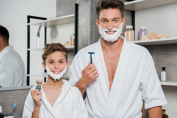 Panoramic shot of father and son with shaving foam on faces holding razor and shaving brush — Stock Photo