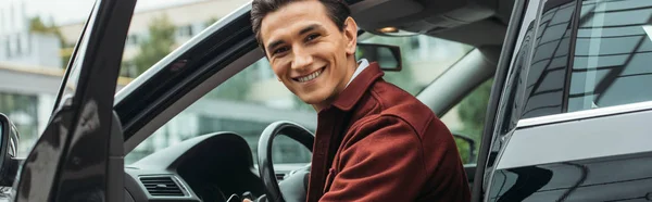 Taxi conductor sentado en el coche con la puerta abierta y sonriendo a la cámara, plano panorámico - foto de stock