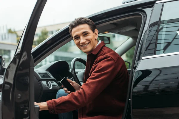 Taxista sonriente sentado en el coche con la puerta abierta y sosteniendo el teléfono inteligente - foto de stock