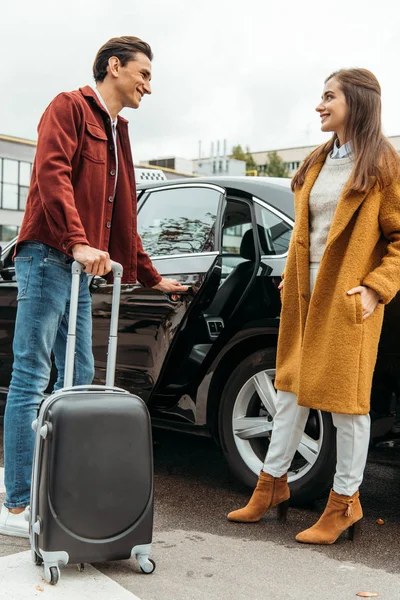 Taxi driver holding wheeled suitcase and smiling to woman by car — Stock Photo