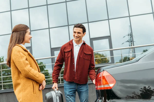 Taxi driver holding suitcase and smiling to passenger by car trunk — Stock Photo