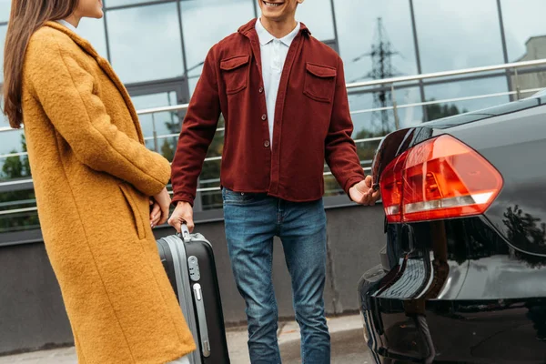 Обрезанный вид водителя такси, держащего чемодан и улыбающегося пассажиру на багажнике автомобиля — стоковое фото