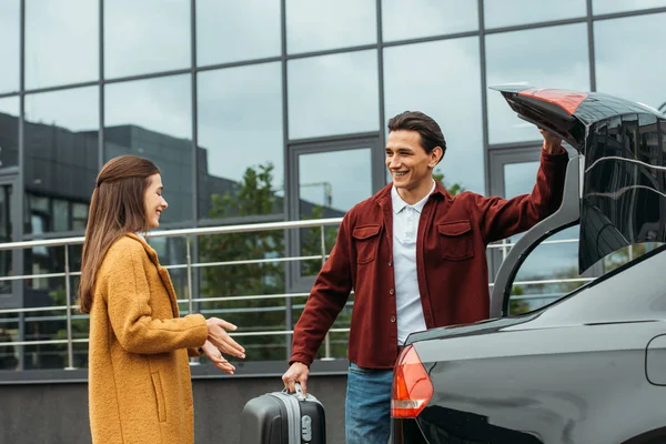 Taxi driver holding suitcase and smiling to woman by open car trunk — Stock Photo